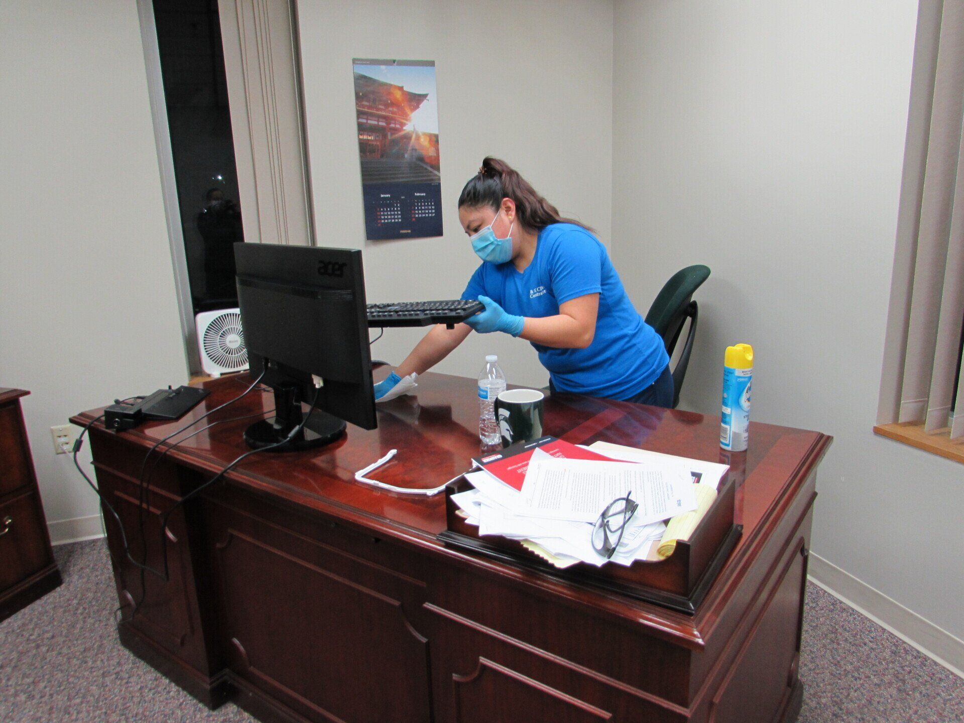 Woman in blue shirt and mask cleaning office desk with computer, keyboard, and cleaning supplies.